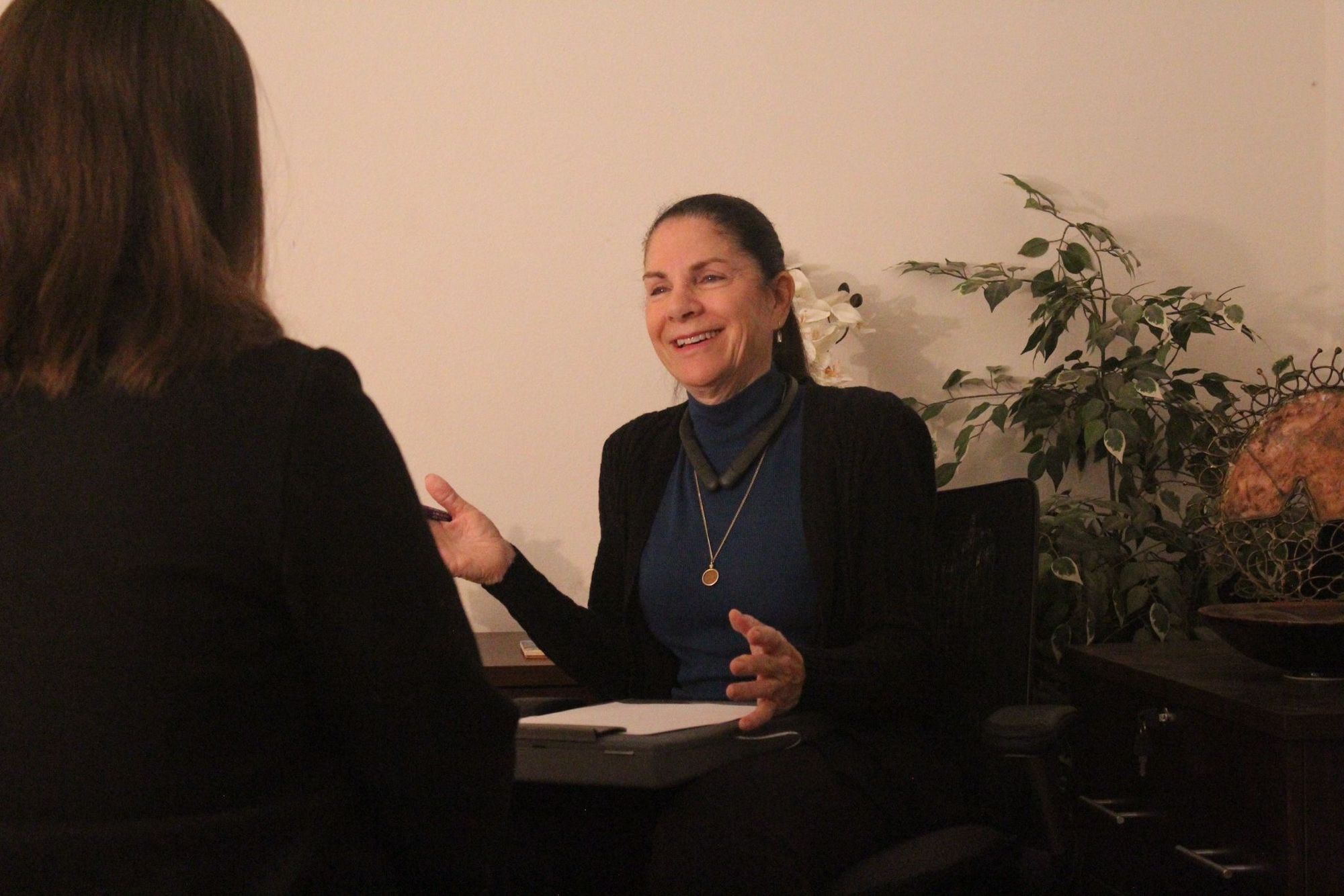 A woman with long hair engages in a conversation, smiling, while sitting at a desk with a notebook. Indoor plants and decor add context to the setting.